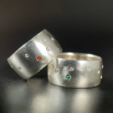 Load image into Gallery viewer, Two silver rings. One is sitting flat on the table and has a scattering of flush-set moissanite stones, with a single bright emerald. The other is leaning on the first at an angle, with a similar random scattering of moissanite and a single orange sapphire.
