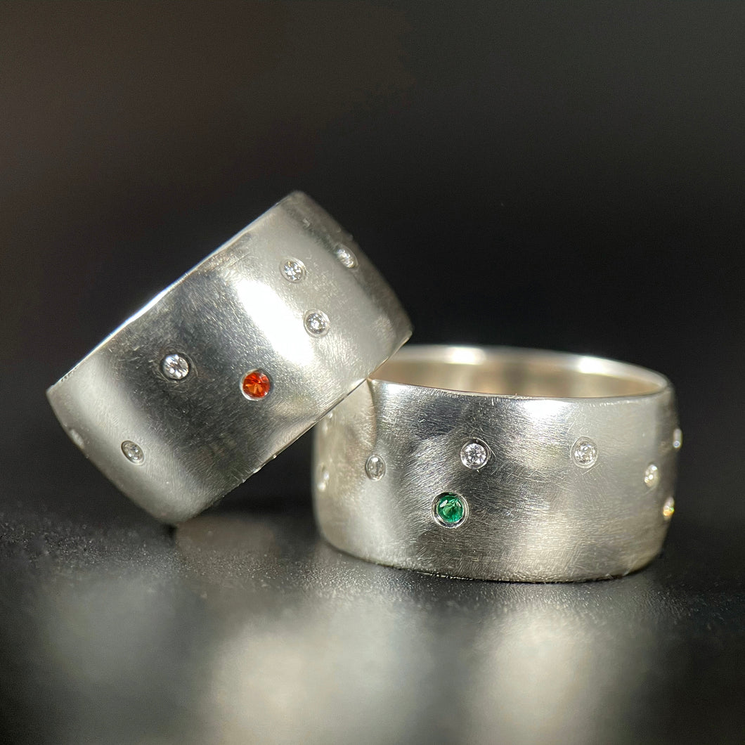 Two silver rings. One is sitting flat on the table and has a scattering of flush-set moissanite stones, with a single bright emerald. The other is leaning on the first at an angle, with a similar random scattering of moissanite and a single orange sapphire.