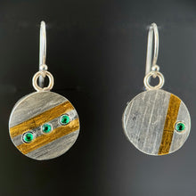 Load image into Gallery viewer, Small round earrings. This side is silver impressed with a diagonal line texture. The earring on the left has a single slanted gold line, with a single emerald near the edge. The other earring has a pair of parallel lines in gold, and between them is a line of 3 small emeralds.
