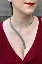 Load image into Gallery viewer, A closeup of a woman wearing a curved silver neckpiece, showing her from her lips down to her bust. The neckpiece curves around the back of her neck and then curves down the centerline of her chest. Grade 2 braille on the piece reads &quot;To love another person is to see the face of God&quot;. The word &quot;God&quot; is done in gold.
