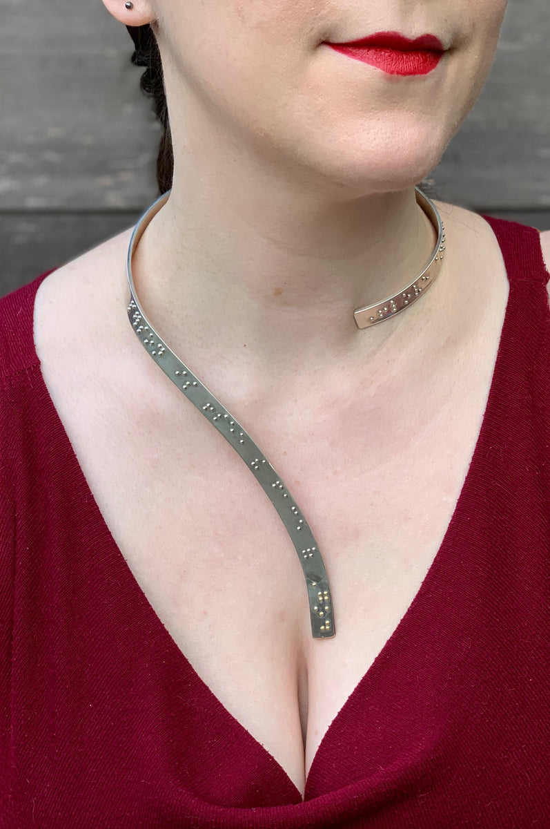 A closeup of a woman wearing a curved silver neckpiece, showing her from her lips down to her bust. The neckpiece curves around the back of her neck and then curves down the centerline of her chest. Grade 2 braille on the piece reads "To love another person is to see the face of God". The word "God" is done in gold.