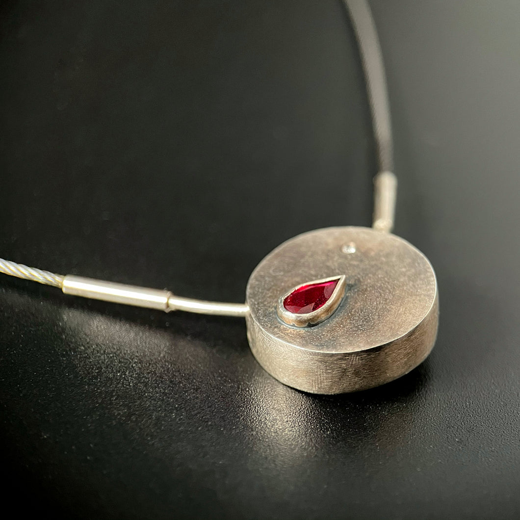 A small round pendant of textured sterling silver. In the upper right is a small round white cubic zirconia, and on the left is a pear-cut lab-grown ruby, the point facing the center of the piece.