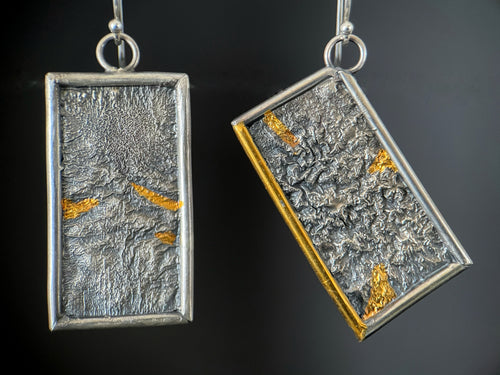 A pair of reversible earrings. These are rectangles framed in silver wire. The one on the left hangs at an angle. Within the frames is rough-textured reticulated silver. In some of the valleys of the silver texture are small triangles of bright gold. On the tilted piece, on piece of the silver wire frame is also gold.